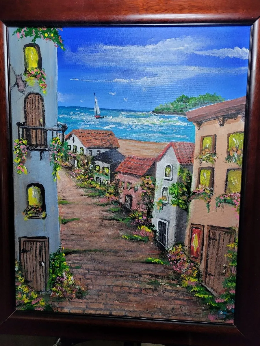 Flower-lined village street with pastel buildings and terracotta roofs overlooking a blue sea and sailboat.