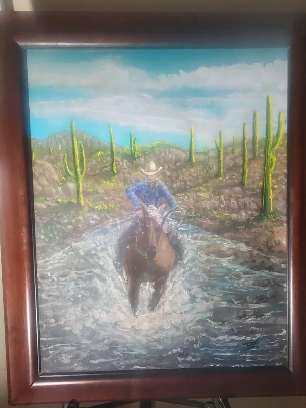 Cowboy in a blue shirt riding a brown horse through splashing water among tall desert cacti.